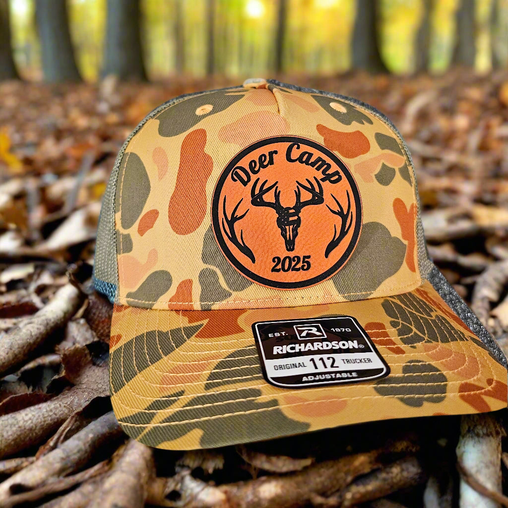 Camouflage trucker hat with 'Deer Camp 2025' patch on a forest floor background