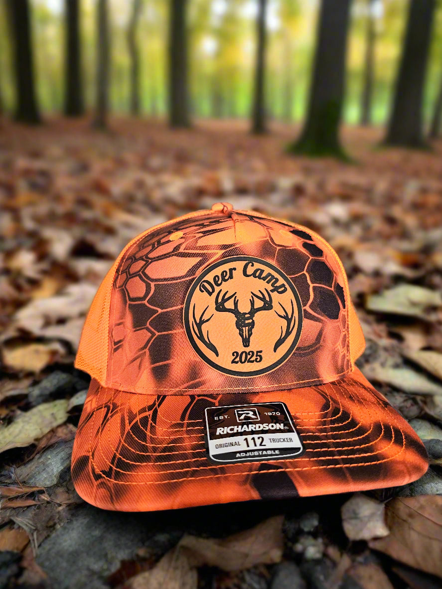 Orange and black cap with 'Deer Camp 2025' logo on a forest floor background