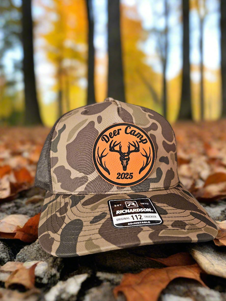 Camouflage cap with 'Deer Camp 2025' logo on a forest floor background