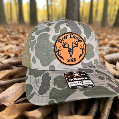 Camouflage cap with 'Deer Camp 2025' patch on a forest background