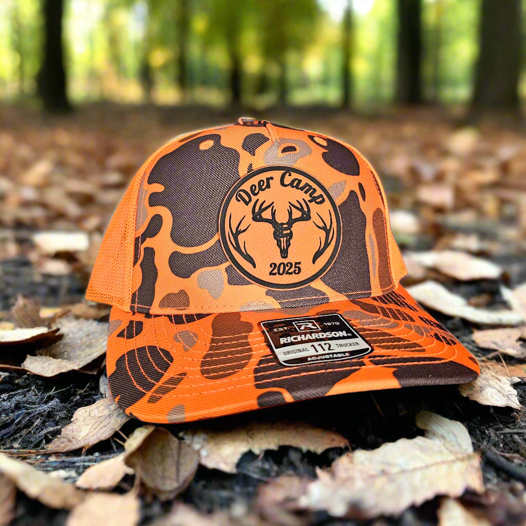 Orange and black camouflage cap with 'Deer Camp 2025' logo on a forest floor background