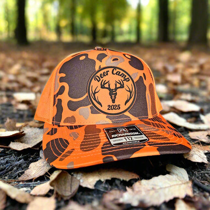 Orange and black camouflage cap with 'Deer Camp 2025' logo on a forest floor background