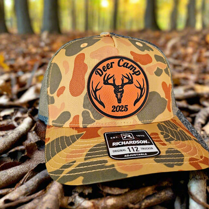 Camouflage trucker hat with 'Deer Camp 2025' patch on a forest floor background